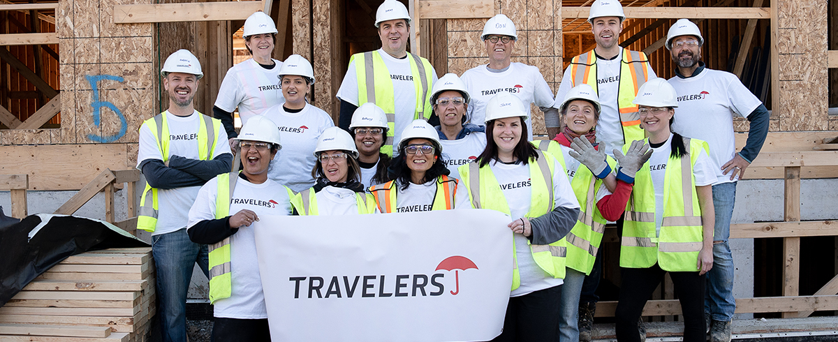 A group of people in white shirts and hard hats hold a "Travelers" banner and pose in front of a construction site